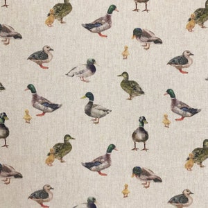 Puede incluir: Una tela beige con un patrón repetido de patos y patitos. Los patos son de varios tonos de marrón, verde y azul. Los patitos son amarillos.