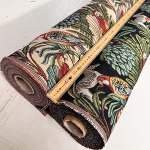 Può includere: Due rotoli di tessuto con un motivo di pappagalli e fogliame tropicale. Un rotolo ha uno sfondo nero, l'altro uno sfondo crema. Un righello di legno è appoggiato sui rotoli di tessuto. Il tessuto presenta pappagalli rossi, blu, gialli e verdi.