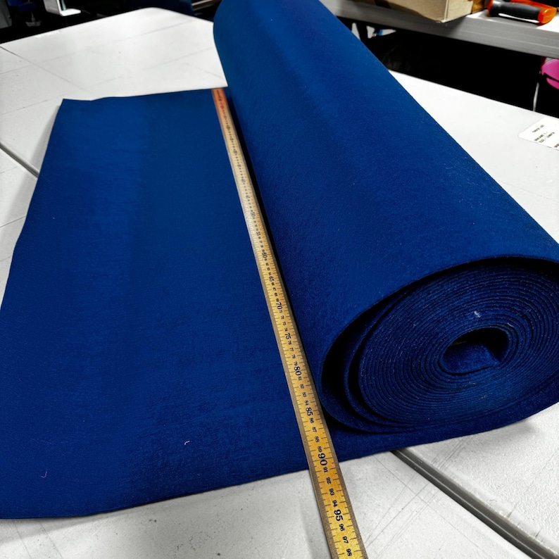 Puede incluir: Un rollo de tela de fieltro azul oscuro, que mide 152,4 cm de largo, se coloca sobre una superficie blanca. Una regla de madera se coloca junto a la tela.