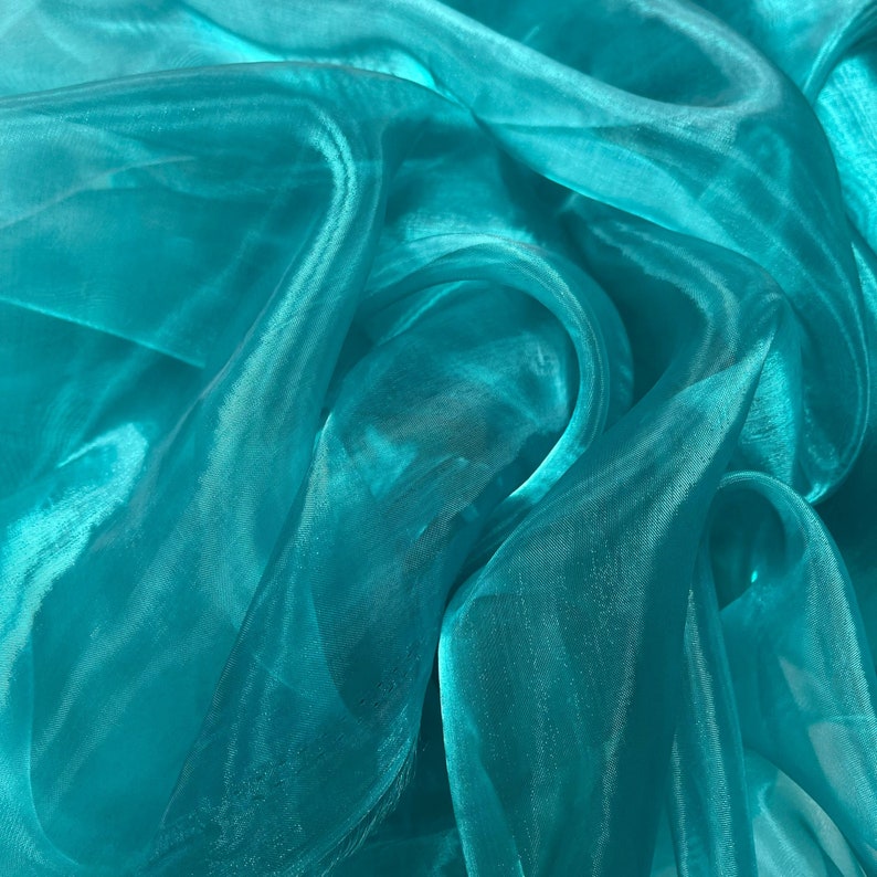 May include: Teal sheer fabric with a soft, flowing texture. The fabric is draped and folded, creating a sense of movement and depth.