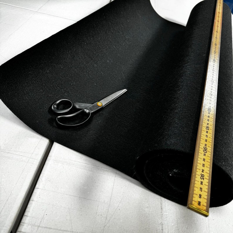 Puede incluir: Un rollo de tela de fieltro negro con una regla de madera marcada con n&uacute;meros del 84 al 99. Un par de tijeras plateadas est&aacute; sobre la tela.