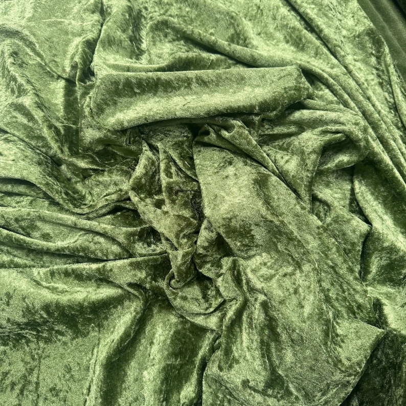 Puede incluir: Una pieza de tela de terciopelo arrugado en un color verde oliva intenso. El material presenta una textura irregular y distintiva con reflejos que captan la luz y sombras profundas creadas por los pliegues y drapeados sobre la superficie.