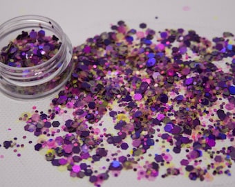 May include: A small clear jar filled with purple, pink, and yellow glitter. The glitter is scattered on a white surface.