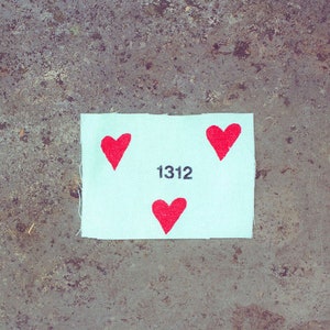 Peut inclure: Un patch en tissu blanc avec trois cœurs rouges et le nombre 1312 imprimé en noir.