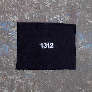 Può includere: Un cerotto quadrato nero con il numero 1312 stampato in bianco su una superficie grigia testurizzata.