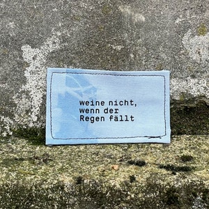 May include: A small, rectangular patch with the German text "weine nicht, wenn der Regen fällt" (don't cry when the rain falls) in black lettering. The patch has a light blue background with a sky-like design and a black stitched border.