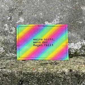 Könnte beinhalten: Ein regenbogenfarbener Stoff-Patch mit dem Text "weine nicht, wenn der Regen fällt" in Schwarz.