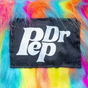 Könnte beinhalten: Ein schwarzer Patch mit weißem Text, der "Dr Pepper" auf einem verschwommenen Hintergrund in Regenbogenfarben lautet.