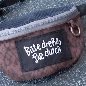 May include: A brown and grey fanny pack with a black adjustable strap. The fanny pack has a zipper closure and a patch with the text "Bitte drehen Sie durch" in white.