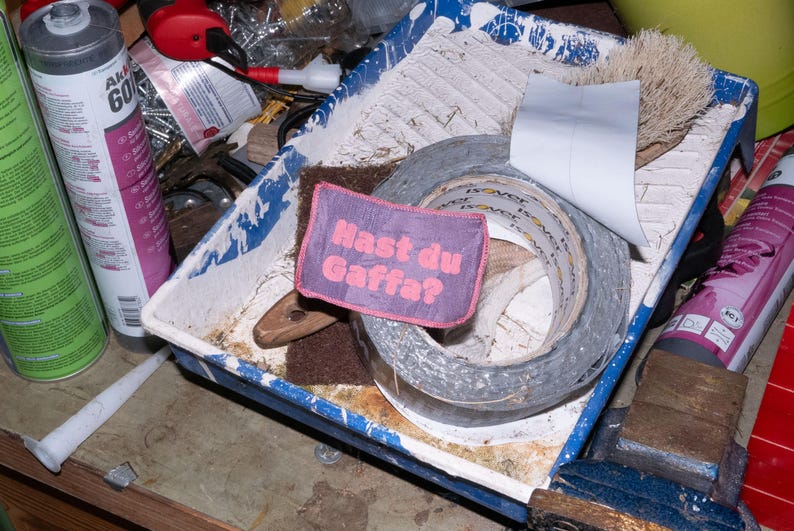 Peut inclure: Un &eacute;cusson rectangulaire en tissu violet avec le texte brod&eacute; en rose "Hast du Gaffa ?" pos&eacute; sur un rouleau de ruban adh&eacute;sif gris dans un bac &agrave; peinture blanc et bleu, entour&eacute; de divers outils de construction et de cartouches.