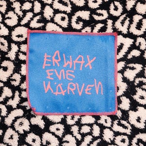 Könnte beinhalten: Ein quadratischer blauer Stoffaufnäher mit rot vernähten Kanten und dem rosa Schriftzug ERWAX EVE KARVEN, zentriert auf einem schwarzen und beigen Textilhintergrund mit Leopardenmuster. Der Aufnäher hat eine glatte Textur und ist aufgenäht.