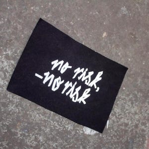 Könnte beinhalten: Schwarzer Stoff-Patch mit weißem Text, der "No risk, no risk" lautet.