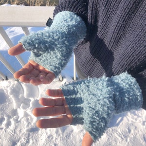 Könnte beinhalten: Ein Paar hellblaue Fingerlose Handschuhe aus einem flauschigen, strukturierten Stoff. Die Handschuhe werden von einer Person mit ausgestreckten Händen getragen.