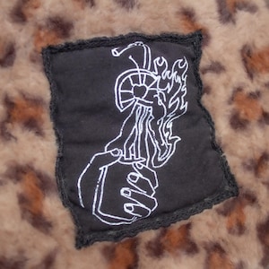 Könnte beinhalten: Ein schwarzer Aufnäher mit einer weißen Grafik einer Hand, die ein Feuerzeug mit Flammen hält, vor einem Leopardenmuster-Hintergrund. Der Aufnäher hat einen schwarzen Rand und ist auf einen weichen, strukturierten Stoff genäht.