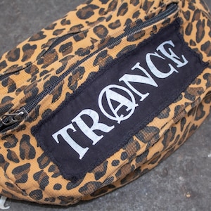 Peut inclure: Une banane de couleur léopard marron et noir avec un patch noir sur lequel est inscrit "TRANCE" en lettres blanches avec un cercle et un "A" à l'intérieur.