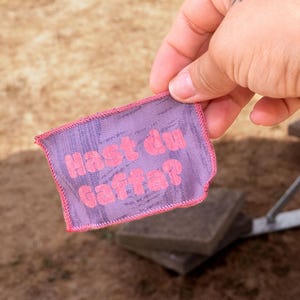 Peut inclure: Un &eacute;cusson rectangulaire en tissu violet avec une surface textur&eacute;e et des bords cousus en rose. Le texte "Hast du Gaffa?" est imprim&eacute; en lettres roses vives et grasses au centre de l'&eacute;cusson, tenu devant un arri&egrave;re-plan ext&eacute;rieur flou.