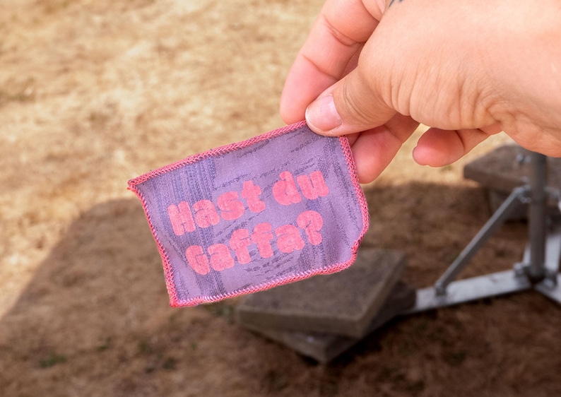 Peut inclure: Un &eacute;cusson rectangulaire en tissu violet avec une surface textur&eacute;e et des bords cousus en rose. Le texte "Hast du Gaffa?" est imprim&eacute; en lettres roses vives et grasses au centre de l'&eacute;cusson, tenu devant un arri&egrave;re-plan ext&eacute;rieur flou.