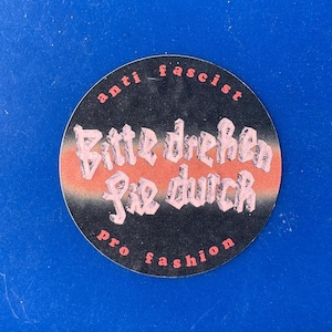 May include: A circular sticker with a black backdrop, featuring the pink text "anti fascist" and "pro fashion". The sticker displays the German phrase "Bitte drehen Sie durch" in a decorative font, set against an orange and pink stripe.