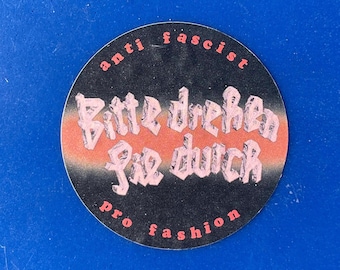 May include: A round sticker with a black background and the words "anti fascist" and "pro fashion" in pink. The sticker has the German phrase "Bitte drehen Sie durch" in a stylized font, with an orange and pink stripe.