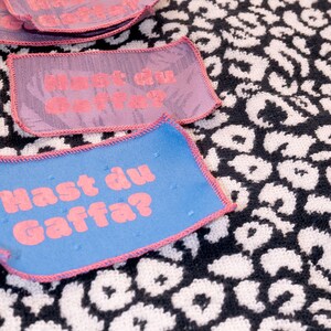 Peut inclure: Des &eacute;cussons rectangulaires en tissu avec des bords cousus roses et le texte "Hast du Gaffa?" en lettres roses. Les &eacute;cussons sont disponibles en bleu et violet, et sont pr&eacute;sent&eacute;s sur un fond textile &agrave; motif l&eacute;opard noir et blanc.