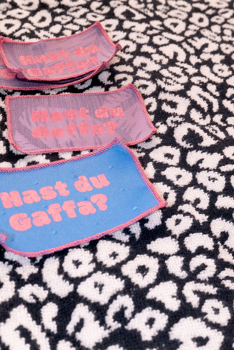 Peut inclure: Des &eacute;cussons rectangulaires en tissu avec des bords cousus roses et le texte "Hast du Gaffa?" en lettres roses. Les &eacute;cussons sont disponibles en bleu et violet, et sont pr&eacute;sent&eacute;s sur un fond textile &agrave; motif l&eacute;opard noir et blanc.