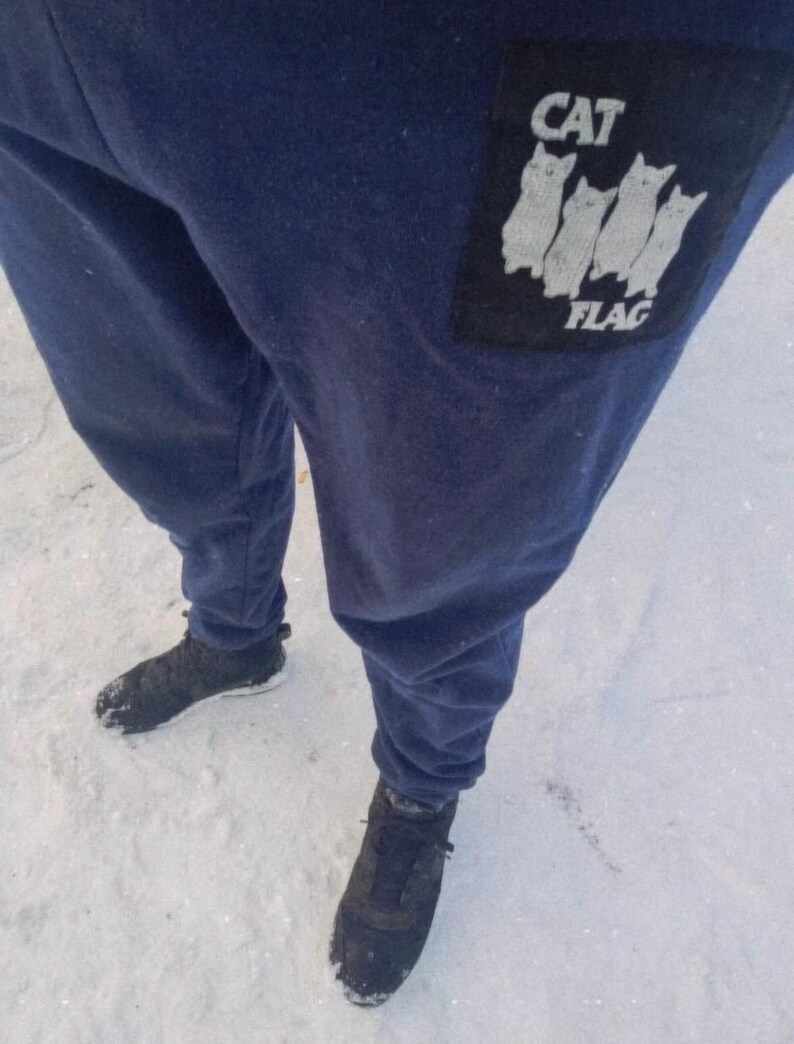 May include: A pair of dark blue sweatpants with a black patch on the left leg. The patch features a white graphic of four cats in a row with the text "CAT FLAG" underneath.