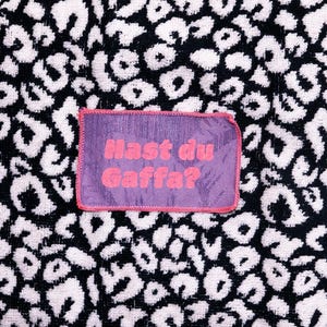 Peut inclure: Un &eacute;cusson rectangulaire en tissu violet avec une bordure cousue rose portant le texte "Hast du Gaffa?" en lettres roses, centr&eacute; sur un fond textile &agrave; motif l&eacute;opard noir et blanc avec un aspect textur&eacute; semblable &agrave; une serviette.