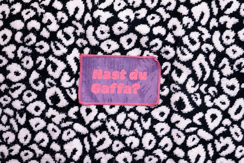 Peut inclure: Un &eacute;cusson rectangulaire en tissu violet avec une bordure cousue rose portant le texte "Hast du Gaffa?" en lettres roses, centr&eacute; sur un fond textile &agrave; motif l&eacute;opard noir et blanc avec un aspect textur&eacute; semblable &agrave; une serviette.