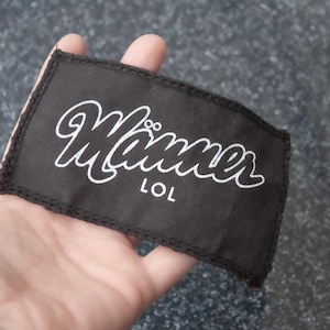 Peut inclure: Patch en tissu noir avec du texte blanc qui dit "Manner LOL".