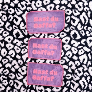 Peut inclure: Trois &eacute;cussons rectangulaires en tissu violet avec des bords cousus en rose. Chaque &eacute;cusson porte le texte "Hast du Gaffa?" imprim&eacute; en rose vif. Les &eacute;cussons pr&eacute;sentent une surface textur&eacute;e et sont dispos&eacute;s verticalement en colonne.