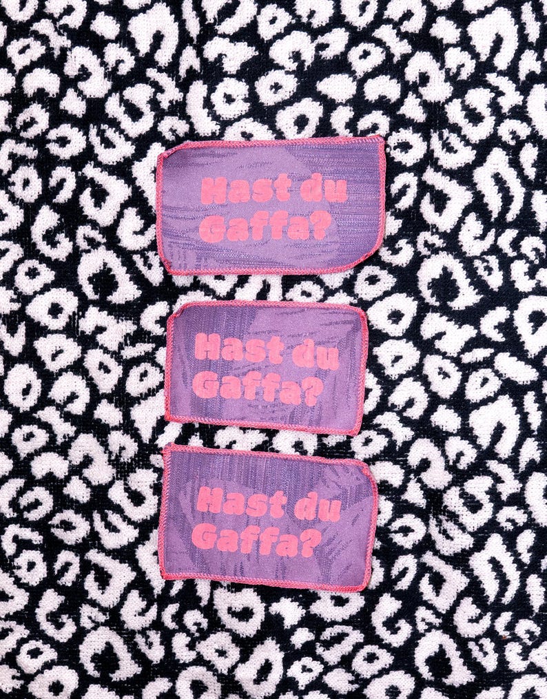Peut inclure: Trois &eacute;cussons rectangulaires en tissu violet avec des bords cousus en rose. Chaque &eacute;cusson porte le texte "Hast du Gaffa?" imprim&eacute; en rose vif. Les &eacute;cussons pr&eacute;sentent une surface textur&eacute;e et sont dispos&eacute;s verticalement en colonne.