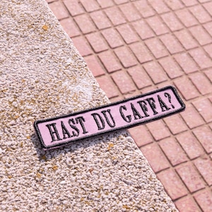 Könnte beinhalten: Ein rechteckiger, rosa Aufnäher mit dem Text "HAST DU GAFFÄ?" in Schwarz, umrandet von einem schwarzen Rand. Der Aufnäher liegt auf einer strukturierten, hellfarbigen Oberfläche mit rotem Backsteinhintergrund.