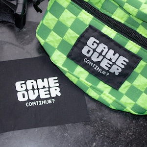 Könnte beinhalten: Eine grüne und schwarze karierte Bauchtasche mit einem schwarzen Patch, auf dem in weißer Schrift "GAME OVER CONTINUE?" steht.