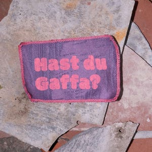 Peut inclure: Un &eacute;cusson rectangulaire en tissu violet avec une bordure cousue rose. Le texte "Hast du Gaffa?" est imprim&eacute; en lettres roses, grasses et arrondies au centre de l'&eacute;cusson, qui pr&eacute;sente un motif textur&eacute; r&eacute;p&eacute;titif et subtil.