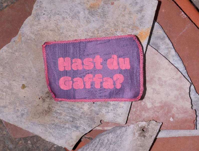 Peut inclure: Un &eacute;cusson rectangulaire en tissu violet avec une bordure cousue rose. Le texte "Hast du Gaffa?" est imprim&eacute; en lettres roses, grasses et arrondies au centre de l'&eacute;cusson, qui pr&eacute;sente un motif textur&eacute; r&eacute;p&eacute;titif et subtil.