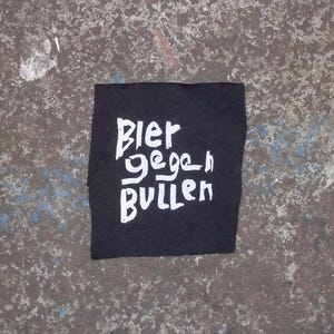 Könnte beinhalten: Schwarzer Stoff-Patch mit weißem Text, der "Bier gegen Bullen" lautet.