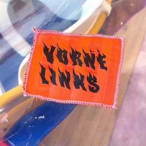 May include: An orange fabric patch with the words "VORNE LINKS" in black, flame-like lettering. The patch has a white stitched border and is set against a blurred background with blue and yellow objects.