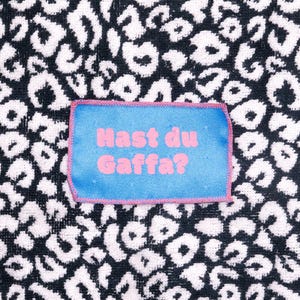 Peut inclure: Un &eacute;cusson rectangulaire en tissu bleu avec des bords cousus roses et le texte rose "Hast du Gaffa?" centr&eacute;. L'&eacute;cusson est plac&eacute; sur un fond en tissu &agrave; motif l&eacute;opard noir et blanc avec une apparence textur&eacute;e semblable &agrave; une serviette.