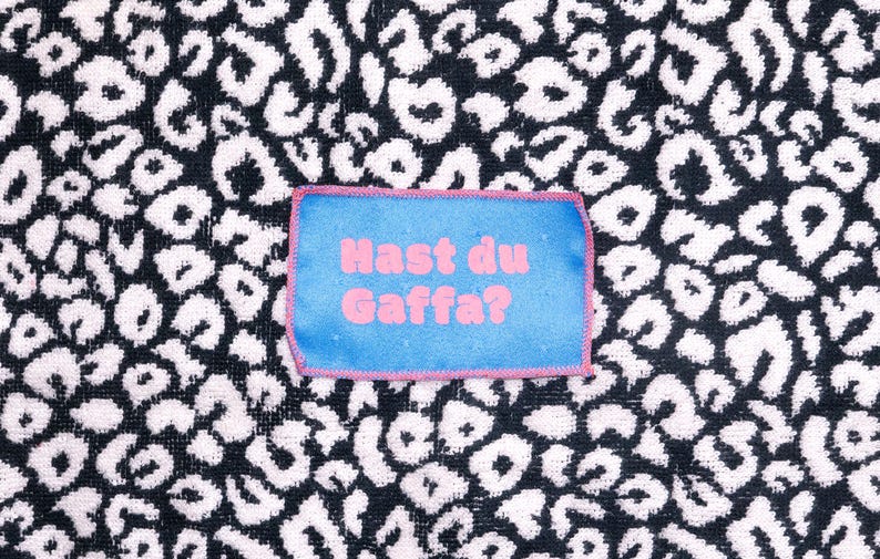 Peut inclure: Un &eacute;cusson rectangulaire en tissu bleu avec des bords cousus roses et le texte rose "Hast du Gaffa?" centr&eacute;. L'&eacute;cusson est plac&eacute; sur un fond en tissu &agrave; motif l&eacute;opard noir et blanc avec une apparence textur&eacute;e semblable &agrave; une serviette.