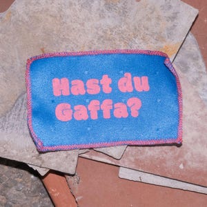 Peut inclure: Un &eacute;cusson rectangulaire en tissu bleu avec des bords cousus roses et le texte rose "Hast du Gaffa?" imprim&eacute; au centre. L'&eacute;cusson pr&eacute;sente un motif &agrave; grille subtil et est pr&eacute;sent&eacute; sur un fond compos&eacute; de plusieurs dalles de pierre superpos&eacute;es.