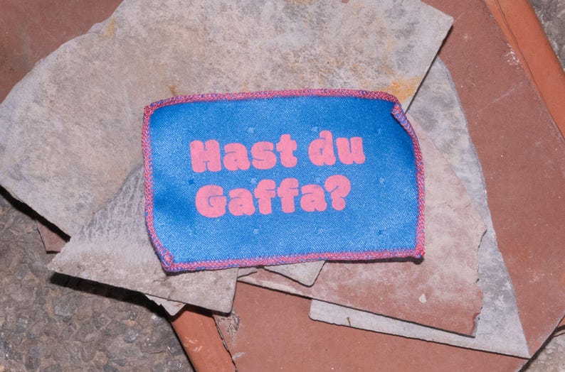 Peut inclure: Un &eacute;cusson rectangulaire en tissu bleu avec des bords cousus roses et le texte rose "Hast du Gaffa?" imprim&eacute; au centre. L'&eacute;cusson pr&eacute;sente un motif &agrave; grille subtil et est pr&eacute;sent&eacute; sur un fond compos&eacute; de plusieurs dalles de pierre superpos&eacute;es.