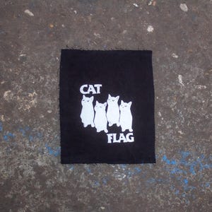 Könnte beinhalten: Schwarzer Stoff-Patch mit weißen gestickten Katzen und dem Text "CAT FLAG".