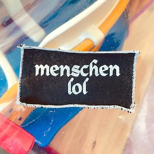 Könnte beinhalten: Ein schwarzer rechteckiger Aufnäher mit weißer gotischer Schrift, die "menschen lol" lautet. Der Aufnäher hat einen weiß genähten Rand. Der Hintergrund ist verschwommen und zeigt Blau, Orange und transparenten Kunststoff.