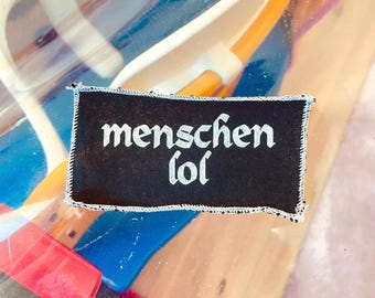 Peut inclure: Un écusson rectangulaire noir avec l'inscription blanche gothique "menschen lol". L'écusson a une bordure blanche cousue. Le fond est flou, montrant du bleu, de l'orange et du plastique transparent.