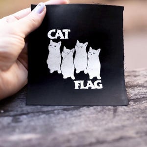 May include: Black fabric patch with white text and a graphic of five cats. The text reads "CAT FLAG".