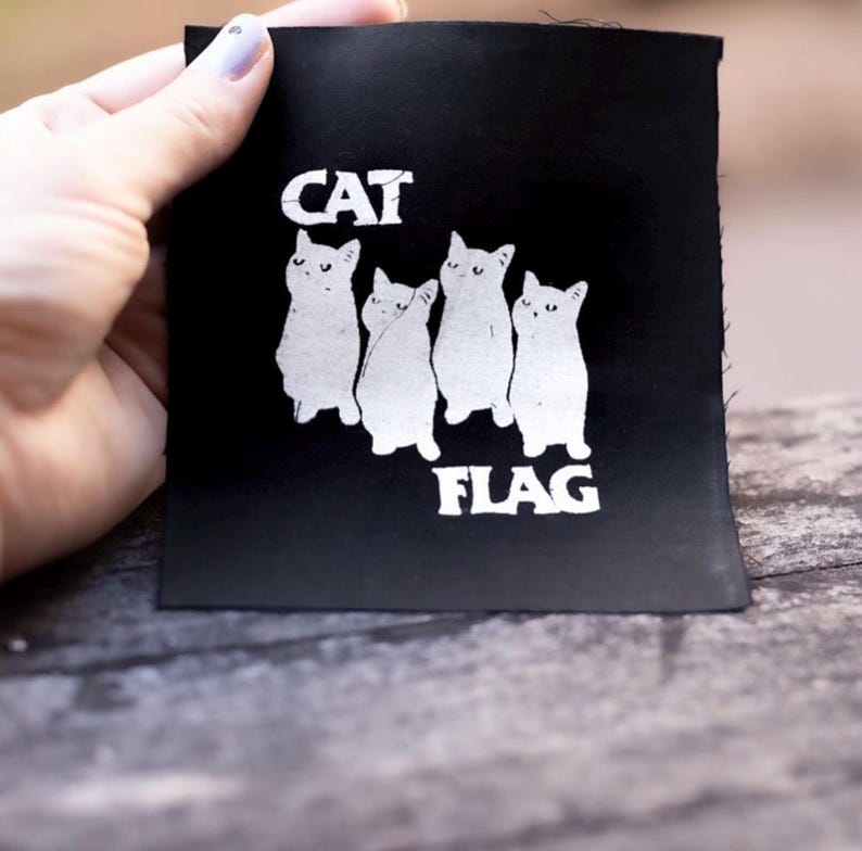 May include: Black fabric patch with white text and a graphic of five cats. The text reads "CAT FLAG".