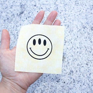 May include: A hand holding a yellow fabric square with a black smiley face with three eyes.