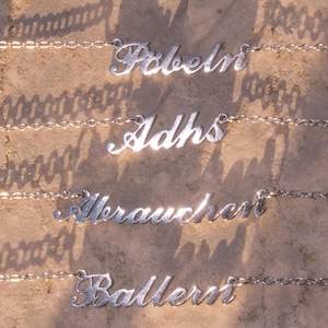 Może przedstawiać: Cztery srebrne naszyjniki z tabliczkami z różnymi imionami w kursywie. Naszyjniki są na łańcuszku. Imiona to: Fabeln, Adhs, Verbraucher i Ballern. Tło to jasnobrązowa powierzchnia.