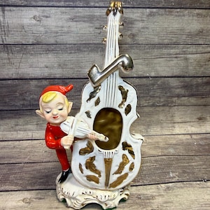 May include: A vintage ceramic figurine featuring a white violin with gold accents and a small elf playing it. The elf wears a red outfit and a pointed hat. The base is white with gold trim.