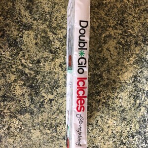 May include: A white package with the words "Double Glo Icicles" in red and black text. The package also has the words "Flameproof" in cursive. The package is on a speckled, brown surface.
